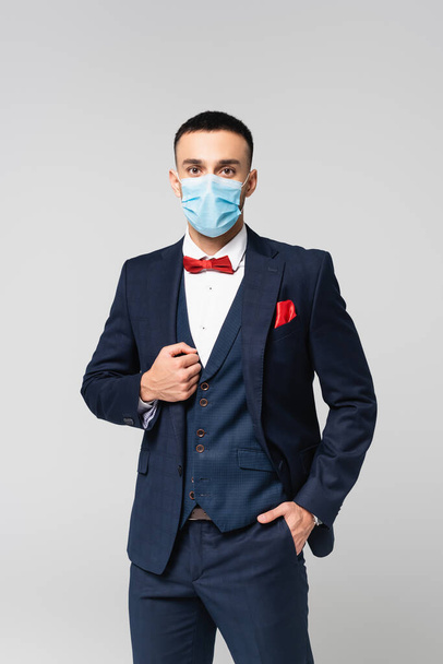κομψός ισπανόφωνος άνδρας με ιατρική μάσκα στέκεται με το χέρι στην τσέπη απομονωμένος στο γκρι - Φωτογραφία, εικόνα