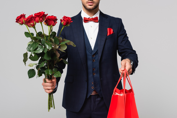 cropped view of man in elegant suit with shopping bags and red roses isolated on grey - Foto, afbeelding