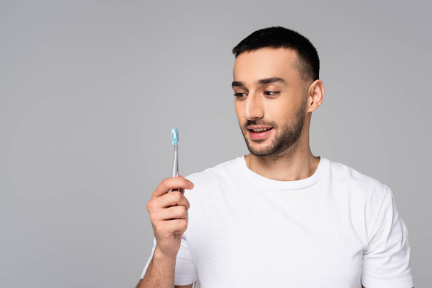 cheerful hispanic man in white t-shirt holding toothbrush isolated on grey - Foto, afbeelding