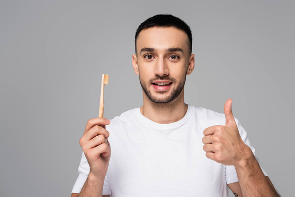 positieve Spaanse man in wit t-shirt met tandenborstel en duim omhoog geisoleerd op grijs - Foto, afbeelding