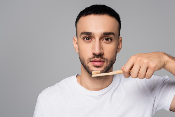 young hispanic man looking at camera while holding toothbrush isolated on grey - Foto, immagini