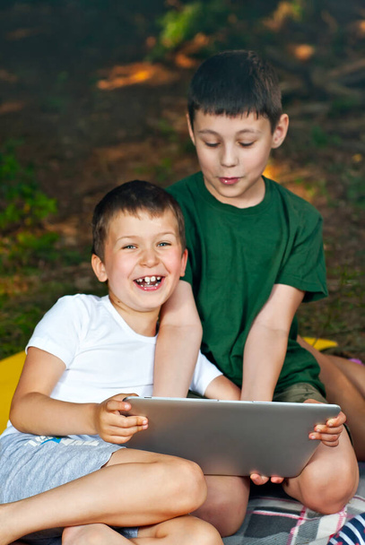 Children play in nature with a laptop. The boys are resting in a tent camp. Children sit on the carpet on the grass in summer. - Φωτογραφία, εικόνα