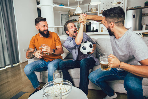 Šťastní přátelé a fotbaloví fanoušci sledující fotbal v televizi a oslavující vítězství doma - Fotografie, Obrázek