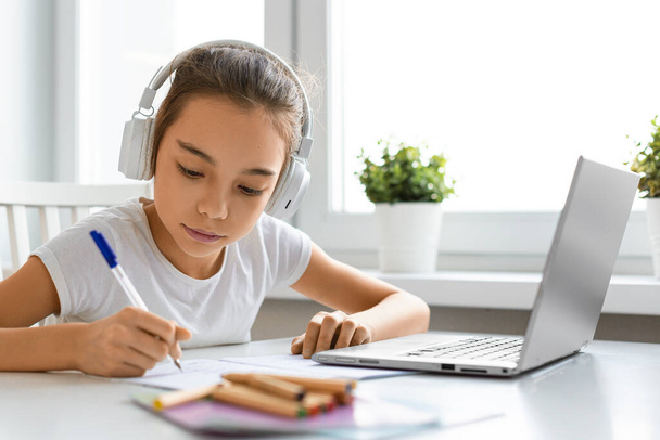 Girl pupil in headphones in front of a laptop monitor makes notes in a notebook durnig online lesson. - Foto, immagini