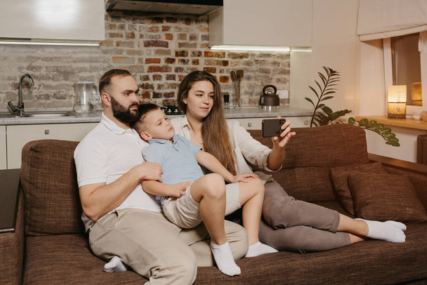 Un padre, un hijo y una madre joven están viendo un video en un teléfono inteligente en el sofá. Una madre está demostrando a su familia noticias en la pantalla del teléfono celular en la noche en casa. - Foto, imagen