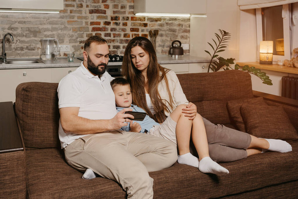 Egy apa, egy fiú és egy fiatal anya néz egy videót egy okostelefonon a kanapén. Egy szakállas apa bemutatja a családi híreket a mobiltelefon képernyőjén este otthon.. - Fotó, kép