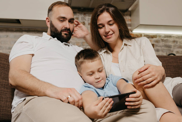 Un padre, un hijo y una madre están viendo videos en el sofá. Un niño está demostrando una caricatura en el teléfono inteligente a la madre y el padre por la noche en casa acogedora.  - Foto, Imagen