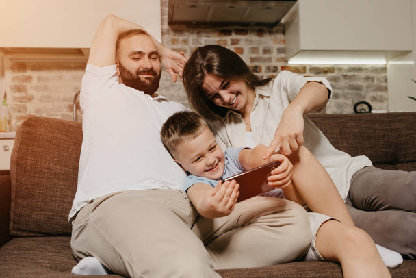 Un padre, un hijo y una madre en el sofá. Un niño feliz está disfrutando del juego en el teléfono inteligente cerca de la madre y el padre sonrientes por la noche en casa acogedora. - Foto, Imagen