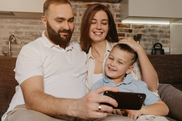 Egy apa, egy fiú és egy anya videóznak a kanapén. Egy boldog férj szakállal bemutatja a műsort az okostelefonon egy mosolygó gyereknek és egy feleségnek este otthon.. - Fotó, kép
