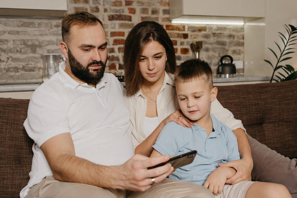 Ένας μπαμπάς, ένας γιος και μια μαμά βλέπουν βίντεο στον καναπέ. Ένας σύζυγος επιδεικνύει την παράσταση στο smartphone στο γελαστό παιδί και τη σύζυγο το βράδυ στο σπίτι.. - Φωτογραφία, εικόνα