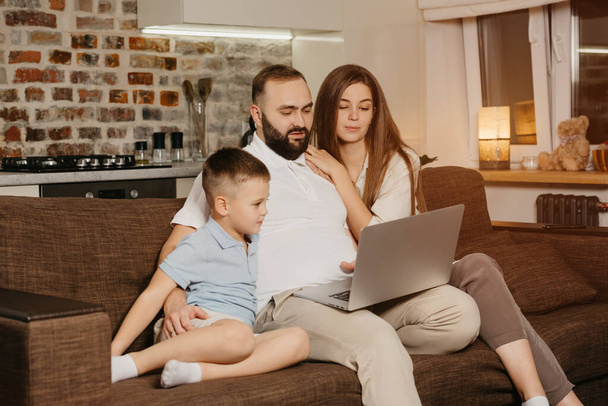 A serious father with a beard is trying to work remotely on a laptop near his son and a curious wife at home. A family at the sofa in the evening. Dad is working online on a computer between relatives - Фото, зображення