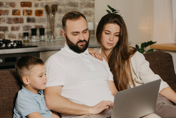 A father with a beard is talking about his achievements at work to a son and a wife at home. A family on the sofa in the evening. Dad is working remotely on a computer between relatives - Photo, Image