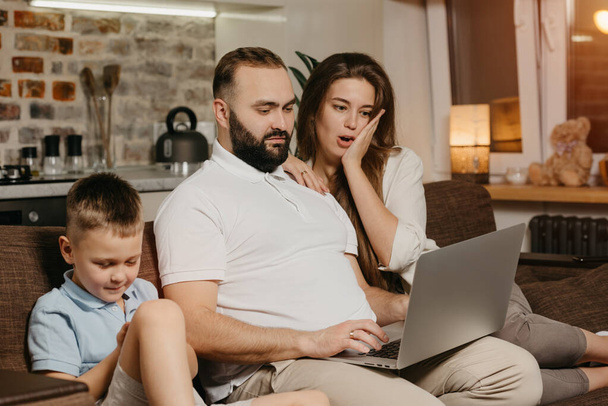 A serious father with a beard is working remotely on a laptop near his son while a wife is surprised at home. A family on the sofa in the evening. Dad is working online on a computer between relatives - Zdjęcie, obraz