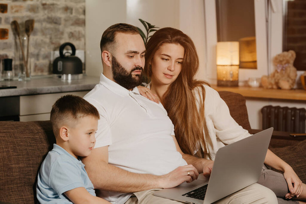 A father with a beard is talking about his achievements at work to his son and wife at home. A young family on the sofa in the evening. Dad is working remotely on a laptop between relatives - Фото, зображення