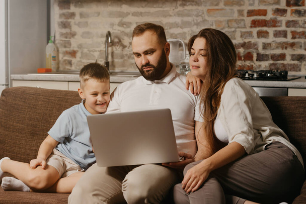 A father with a beard is working remotely on a laptop while his happy son and wife are staring at the screen. Dad is working online on a computer between relatives on the sofa at home in the evening - Photo, Image