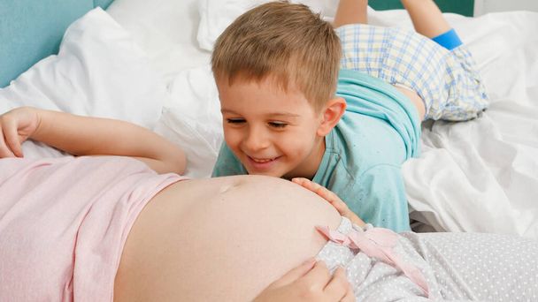 Χαριτωμένο αγοράκι με πιτζάμες να ακούει αγέννητο μωρό σε έγκυο κοιλιά μητέρας ξαπλωμένο στο κρεβάτι. Έννοια της αγάπης των παιδιών και της οικογενειακής ευτυχίας αναμένοντας το μωρό - Φωτογραφία, εικόνα
