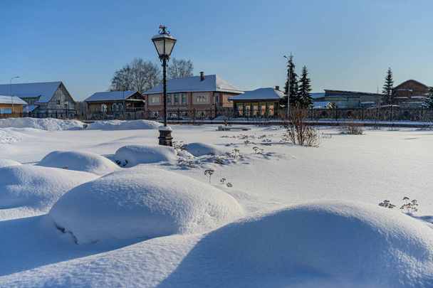 en el parque de la ciudad de Nevyansk (Rusia) en invierno. una gran cantidad de nieve blanca esponjosa en el suelo y las plantas, bolas de nieve formadas. en la distancia una linterna, árboles y casas. cielo azul - Foto, imagen