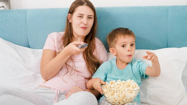 Ευτυχισμένο το μικρό παιδί με τη νεαρή μητέρα βλέποντας ταινία στην τηλεόραση και τρώγοντας ποπ κορν - Φωτογραφία, εικόνα