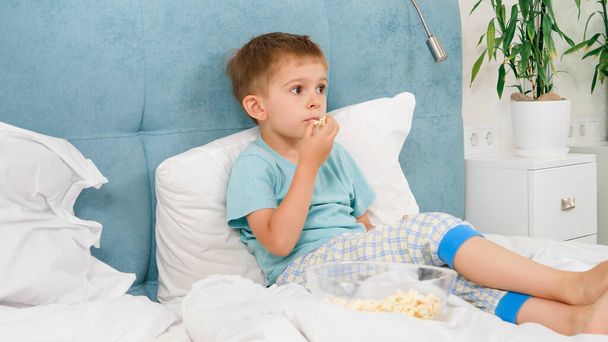 Pequeño niño en pijama acostado en la cama y comiendo palomitas de maíz mientras ve la televisión por la mañana. - Foto, Imagen