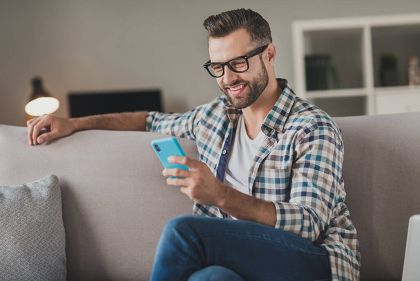 Photo of young cheerful man happy positive smile read browse internet cellphone chat type sms sit couch indoors - Foto, Imagem