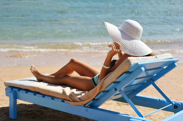 Schöne Frau sonnt sich am Strand eines tropischen Urlaubsortes und genießt den Sommerurlaub. Mädchen mit Hut und Händen auf Sonnenliege am Meer - Foto, Bild