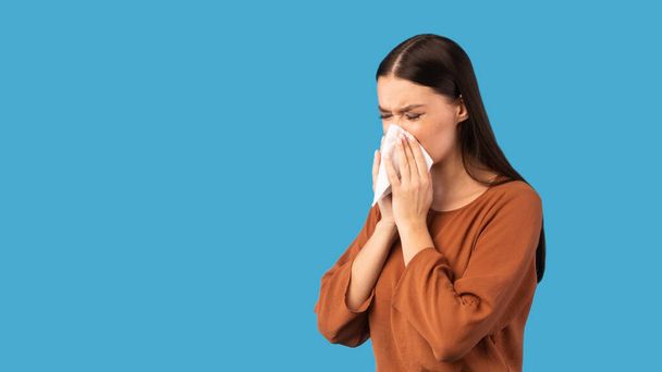 Chora kobieta wieje cieknącym nosem w papierowej tkance, niebieskie tło - Zdjęcie, obraz