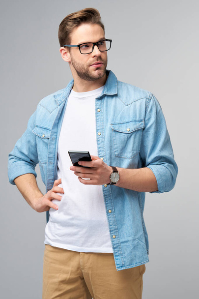 pretty casual man in blue jeans shirt holding his phone standing over studio grey background - Foto, Imagem