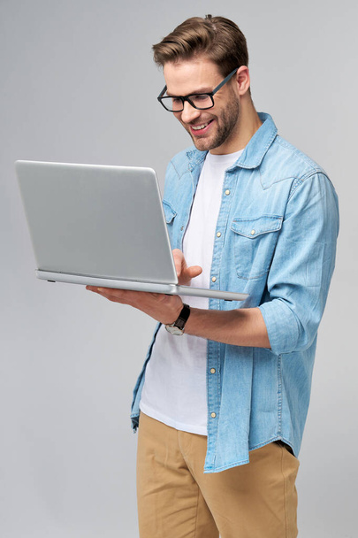 Concentrated young bearded man wearing glasses dressed in jeans shirt using laptop isolated over grey studio background - Zdjęcie, obraz