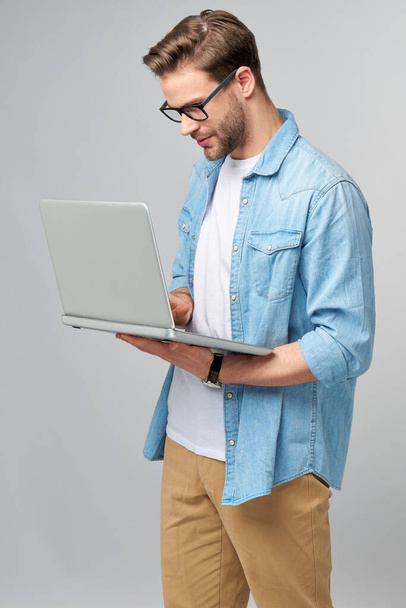 Jeune homme barbu concentré portant des lunettes vêtu d'une chemise en jeans utilisant un ordinateur portable isolé sur fond de studio gris - Photo, image