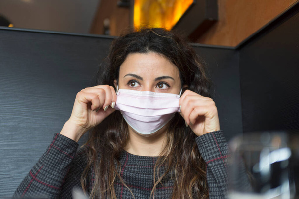 Portret van een jonge vrouw die een medisch masker draagt en aanpast, zittend aan tafel in restaurant - Foto, afbeelding