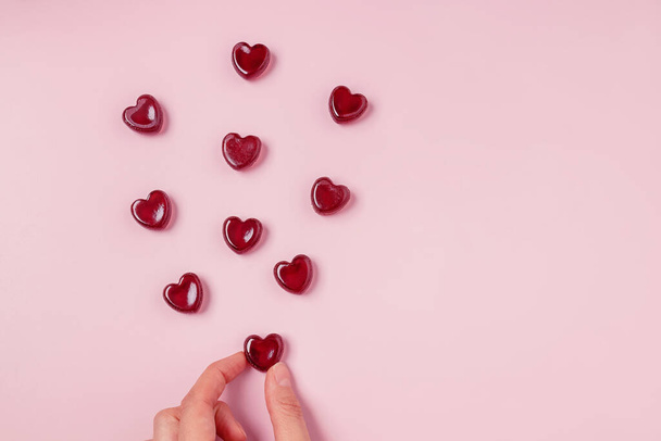 Mano sosteniendo uno de caramelos de jalea roja dispersos de forma de corazón sobre fondo rosa - Foto, imagen