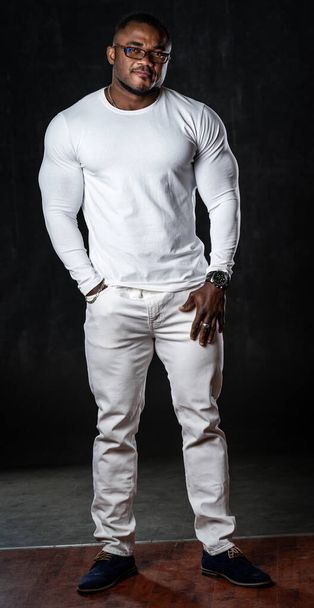 Atleta forte em jeans brancos e camisa casual branca posa para a câmera em fundo preto. Retrato em tamanho real de um fisiculturista afro-americano. - Foto, Imagem