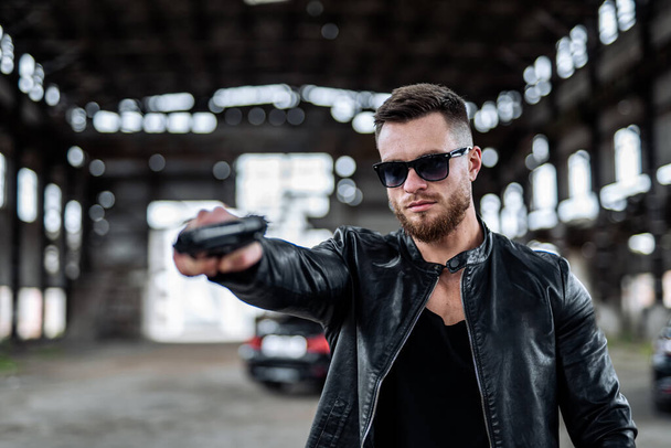 Brutal young man in total black outfit and sunglasses points a gun on some point. Abandoned blurred background of warehouse. - Foto, immagini