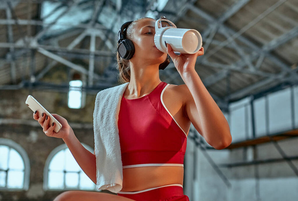 Beautiful woman using smartphone and listening to music in headphones drinks protein shake while exercising in the fitness room. Sports and technology concept. Lifestyle and health theme. - Foto, afbeelding