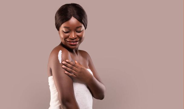 Φροντίδα δέρματος. Όμορφη αφρικάνικη κυρία τυλιγμένη σε πετσέτα εφαρμόζοντας ενυδατική λοσιόν σώματος - Φωτογραφία, εικόνα