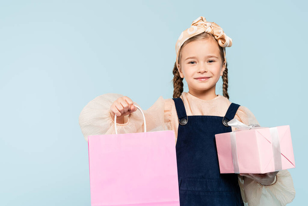 happy kid with pigtails holding shopping bag and present isolated on blue - Foto, immagini
