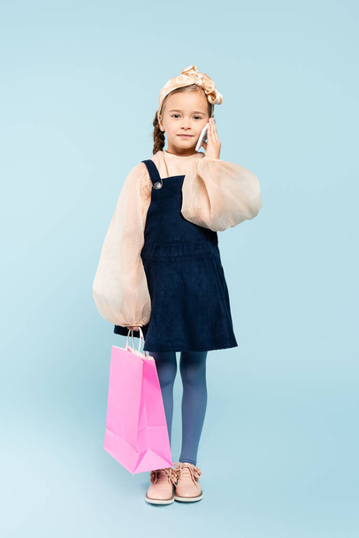 full length of happy kid with pigtails talking on smartphone and holding shopping bag on blue - Valokuva, kuva