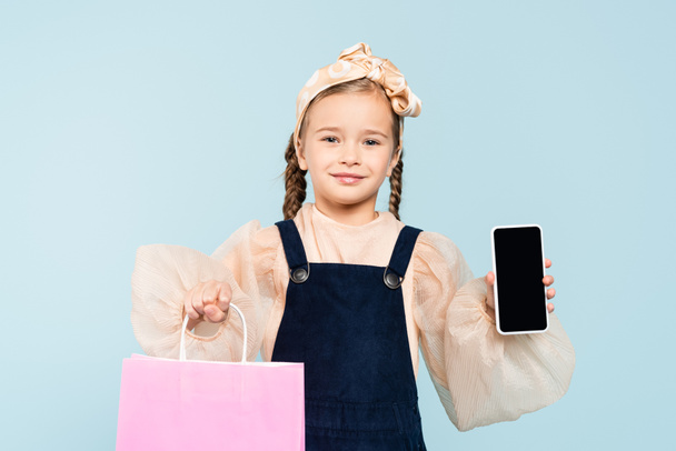 happy kid with pigtails holding smartphone with blank screen and shopping bag isolated on blue - Valokuva, kuva