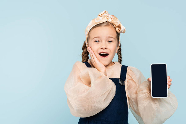 amazed kid with pigtails holding smartphone with blank screen isolated on blue - Photo, image
