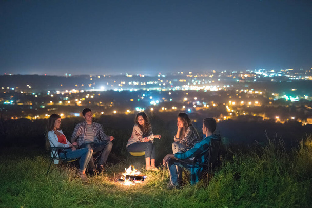 Οι πέντε άνθρωποι κάθονται κοντά σε μια φωτιά στα φώτα της πόλης φόντο. νύχτα - Φωτογραφία, εικόνα