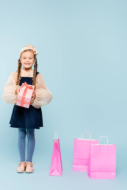 full length of little girl in dress holding present near shopping bags on blue - Фото, изображение