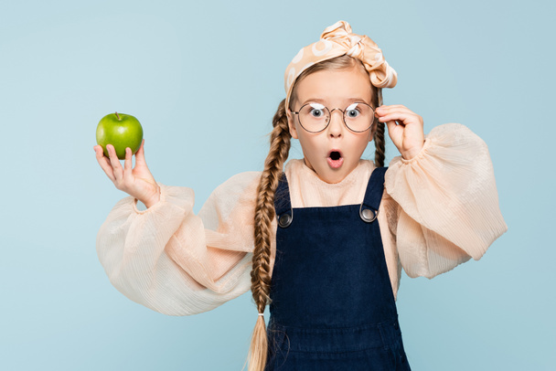 shocked kid adjusting glasses and holding green apple isolated on blue - 写真・画像