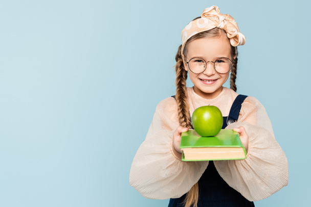 bambino intelligente in bicchieri tenendo libro con mela verde e sorridente isolato su blu - Foto, immagini