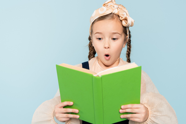 choqué petite fille avec des nattes lecture livre isolé sur bleu - Photo, image