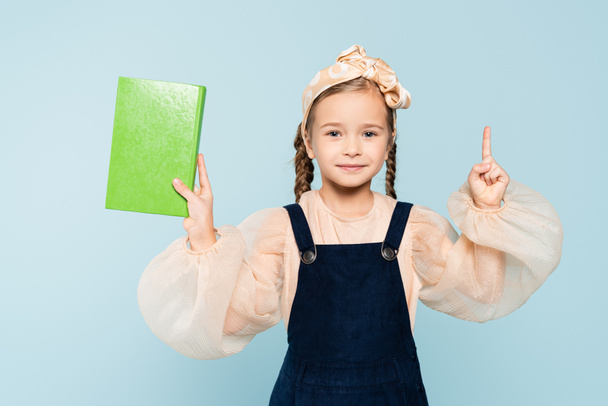 little girl with pigtails holding book and pointing with finger isolated on blue - Фото, изображение