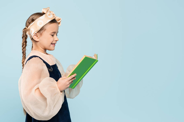 little girl with pigtails smiling while reading book isolated on blue - Фото, изображение