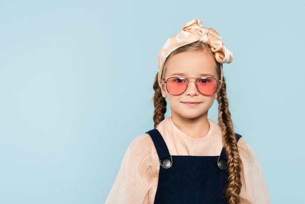 mała dziewczynka w okularach przeciwsłonecznych patrząc na aparat odizolowany na niebiesko - Zdjęcie, obraz