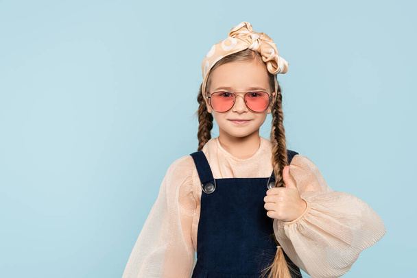 little girl in sunglasses showing thumb up isolated on blue - Foto, afbeelding