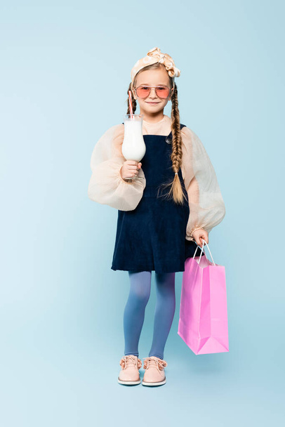 Güneş gözlüklü, elinde bardak, elinde milkshake ve mavi çantalı küçük bir kız çocuğu. - Fotoğraf, Görsel