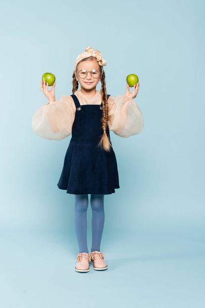 full length of kid in glasses holding green apples on blue - Valokuva, kuva